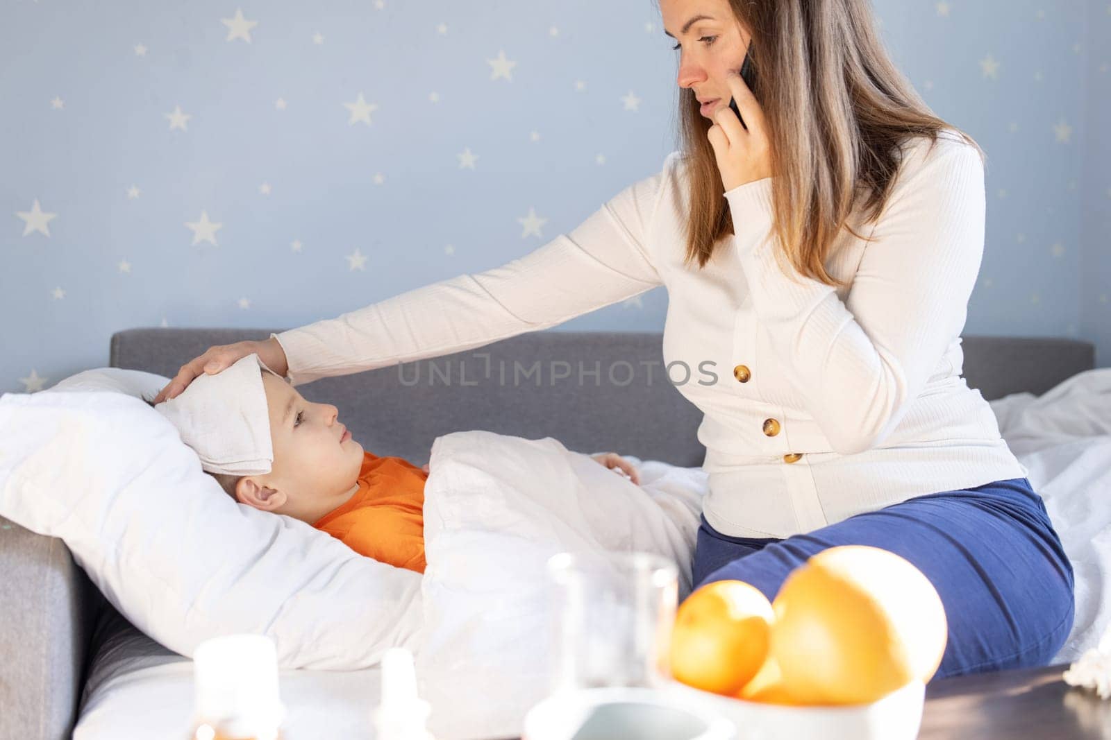 Worried young mother of sick child calling to a doctor on phone. Sick kid feels bad and lying in bed under a blanket at home.