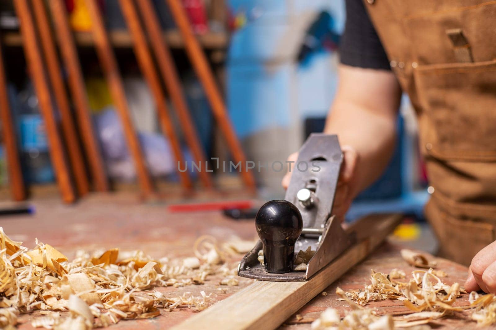 Carpenter in an apron planing a wooden board with a planer, workplace.