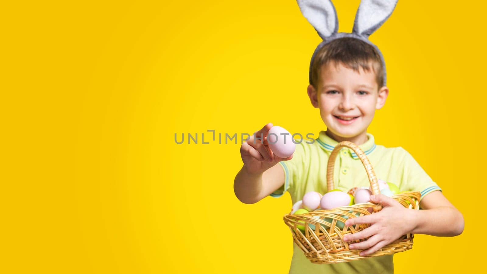 A happy smilling boy with Easter bunny ears holds a basket with painted eggs and shows one of them on a yellow background. Copy space. Easter day.