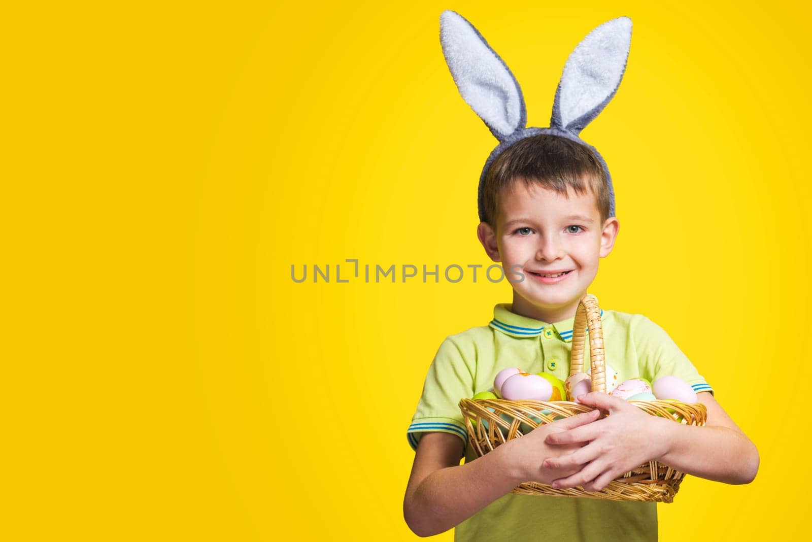 A happy smilling boy wearing Easter bunny ears holds a basket with painted eggs on a yellow background. Copy space. Easter day.