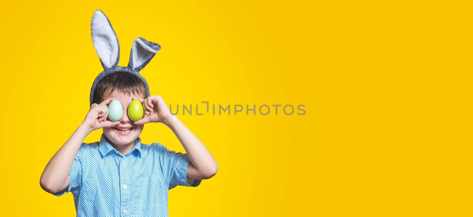 Cute boy with bunny ears keeping colored eggs near eyes on yellow background. Happy child boy with easter eggs and bunny ears on yellow background.