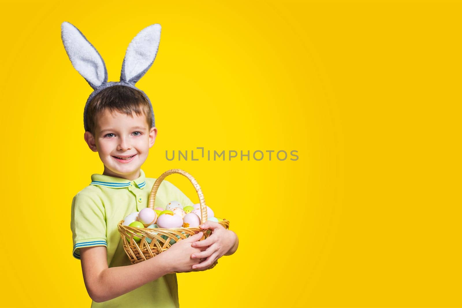 A happy smiling boy wearing Easter bunny ears holds a basket with painted eggs on a yellow background. Copy space. Easter day.