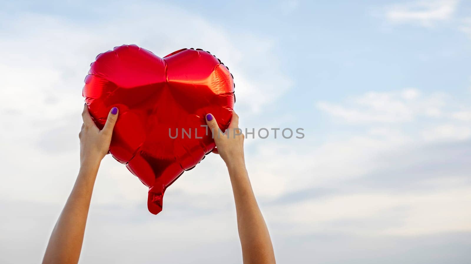 Womans hands with heart shaped balloon on background of sky. Valentine day and romance concept.