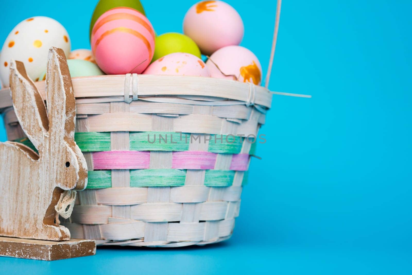 A basket of Easter eggs with toy bunny on blue background. Festive basket with colorful painted eggs and toy wooden rabbit. Easter composition.