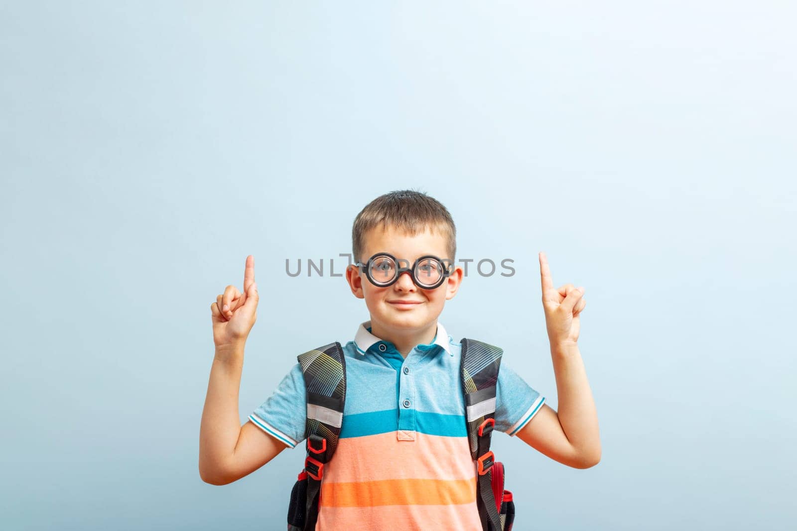 Happy school boy pointing fingers up showing something on blue background in studio. Free space for your text. Back to school.