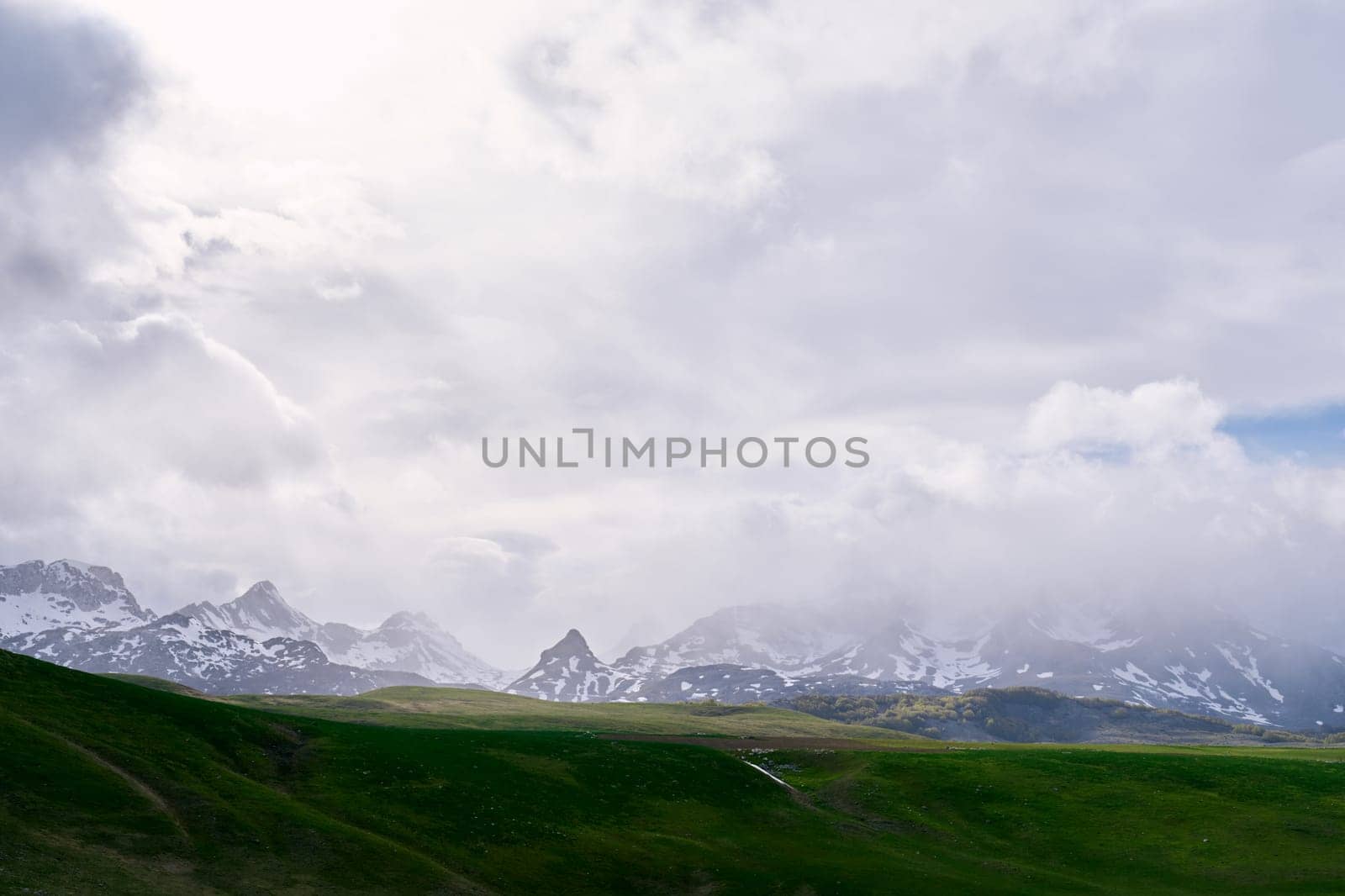 Fog over snow-capped mountains descends onto a green valley. High quality photo