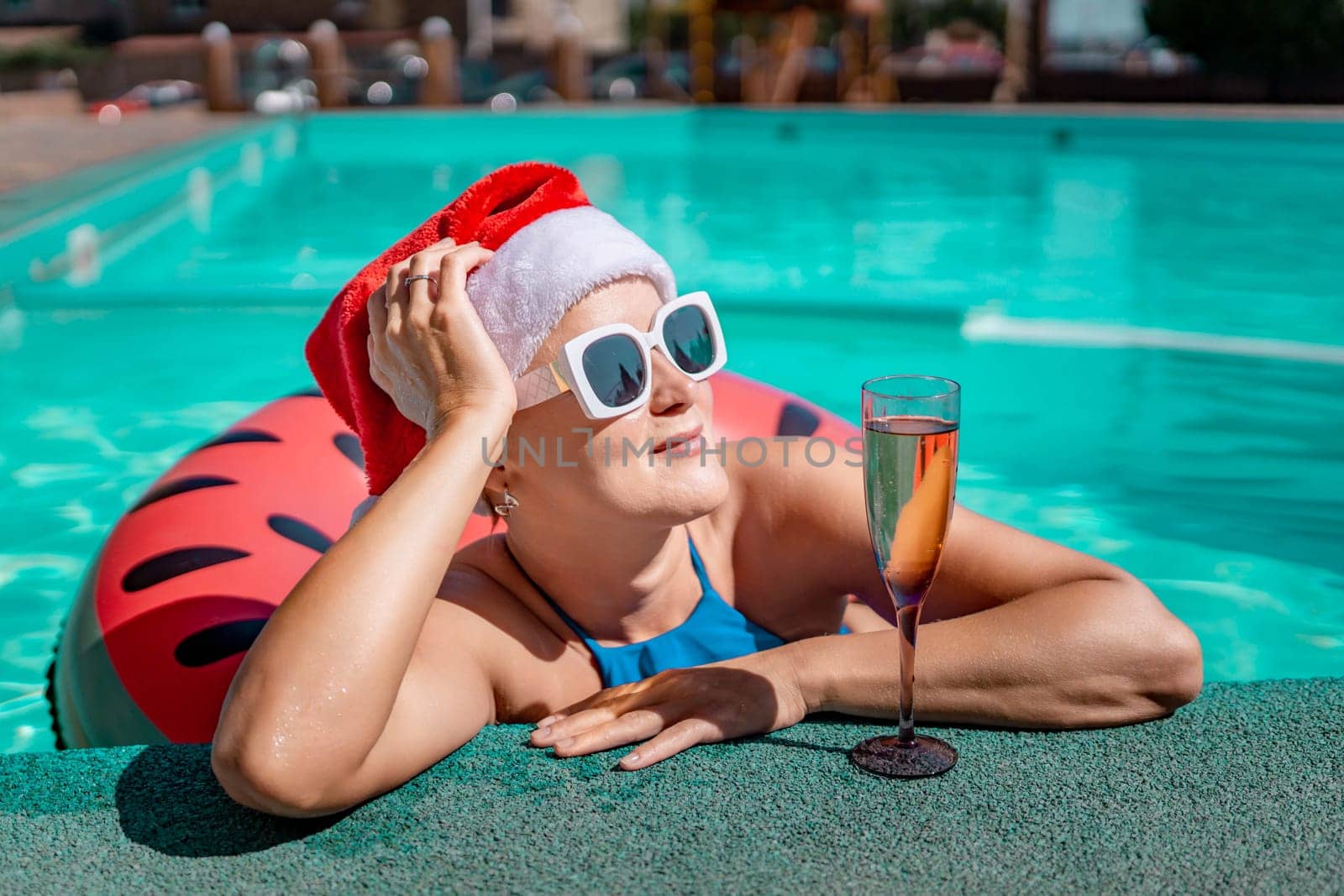 Woman pool Santa hat. A happy woman in a blue bikini, a red and white Santa hat and sunglasses poses near the pool with a glass of champagne standing nearby. Christmas holidays concept. by Matiunina