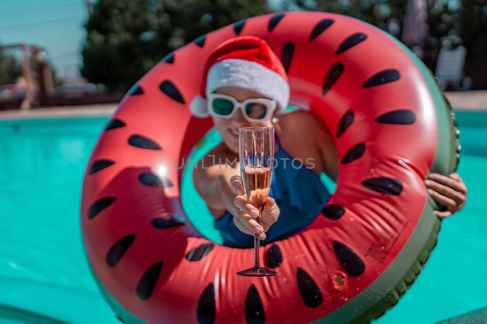 Woman pool Santa hat. A happy woman in a blue bikini, a red and white Santa hat and sunglasses poses near the pool with a glass of champagne standing nearby. Christmas holidays concept
