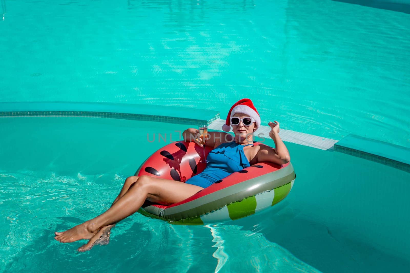 A happy woman in a blue bikini, a red and white Santa hat and sunglasses poses in the pool in an inflatable circle with a watermelon pattern, holding a glass of champagne in her hands. Christmas holidays concept