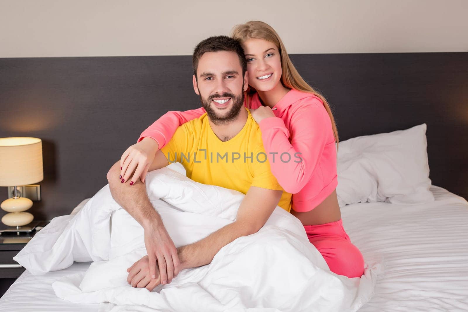 Young lovely couple lying and have fun in a bed, happy smile looking at camera, cover under blanket. Family, bedtime and happiness concept