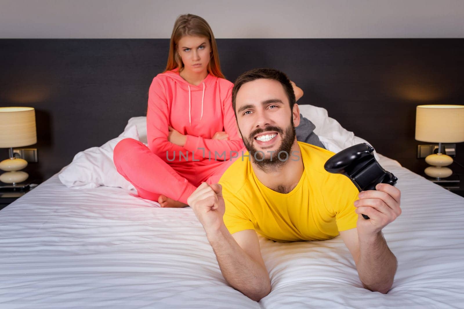 Young couple having playing videogames in bed. Man playing video game, upset woman on background.