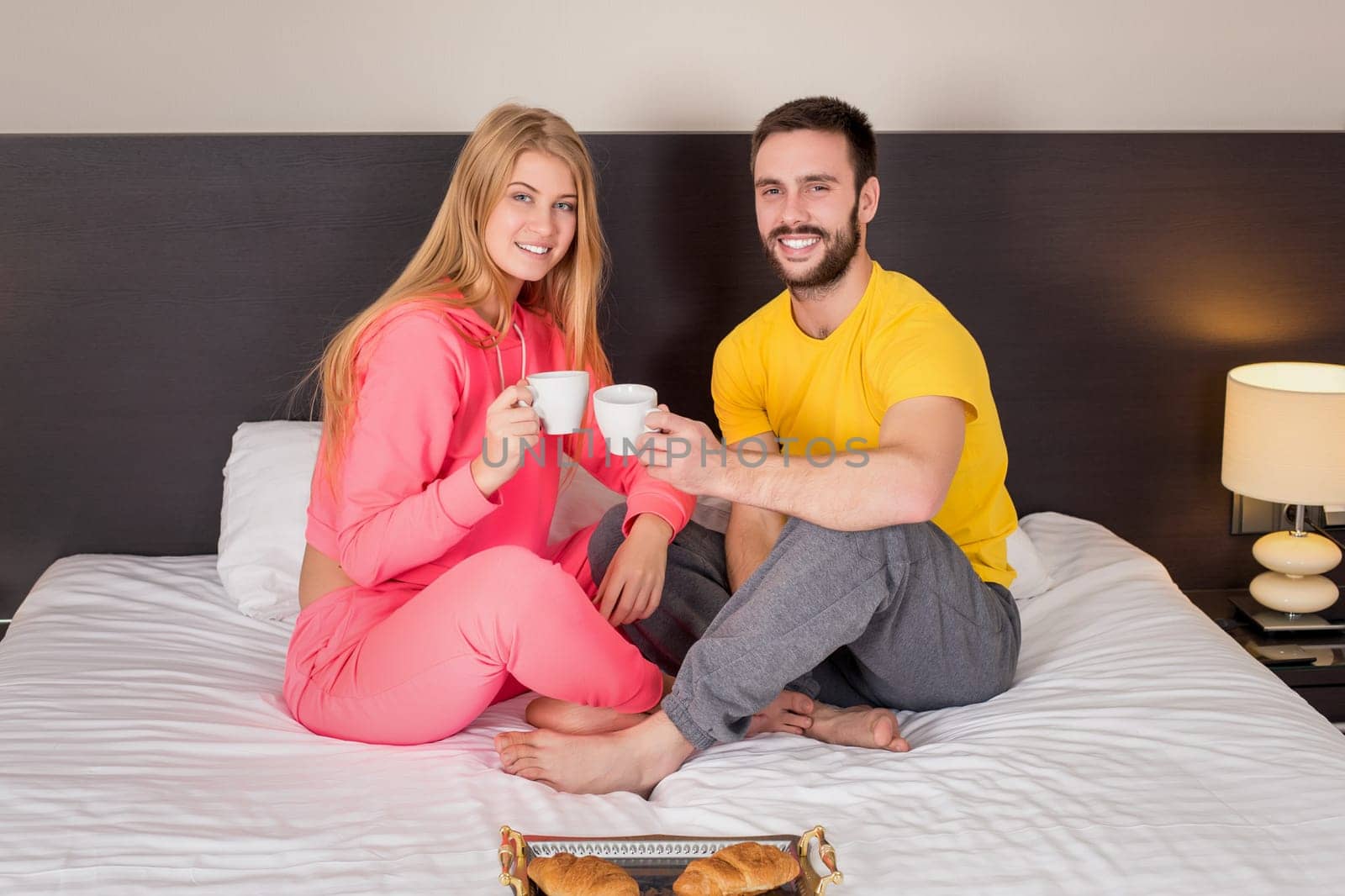 Happy young couple having breakfast tray on bed at home in bedroom. Young couple drink coffee in bed, happy smile woman man lovers