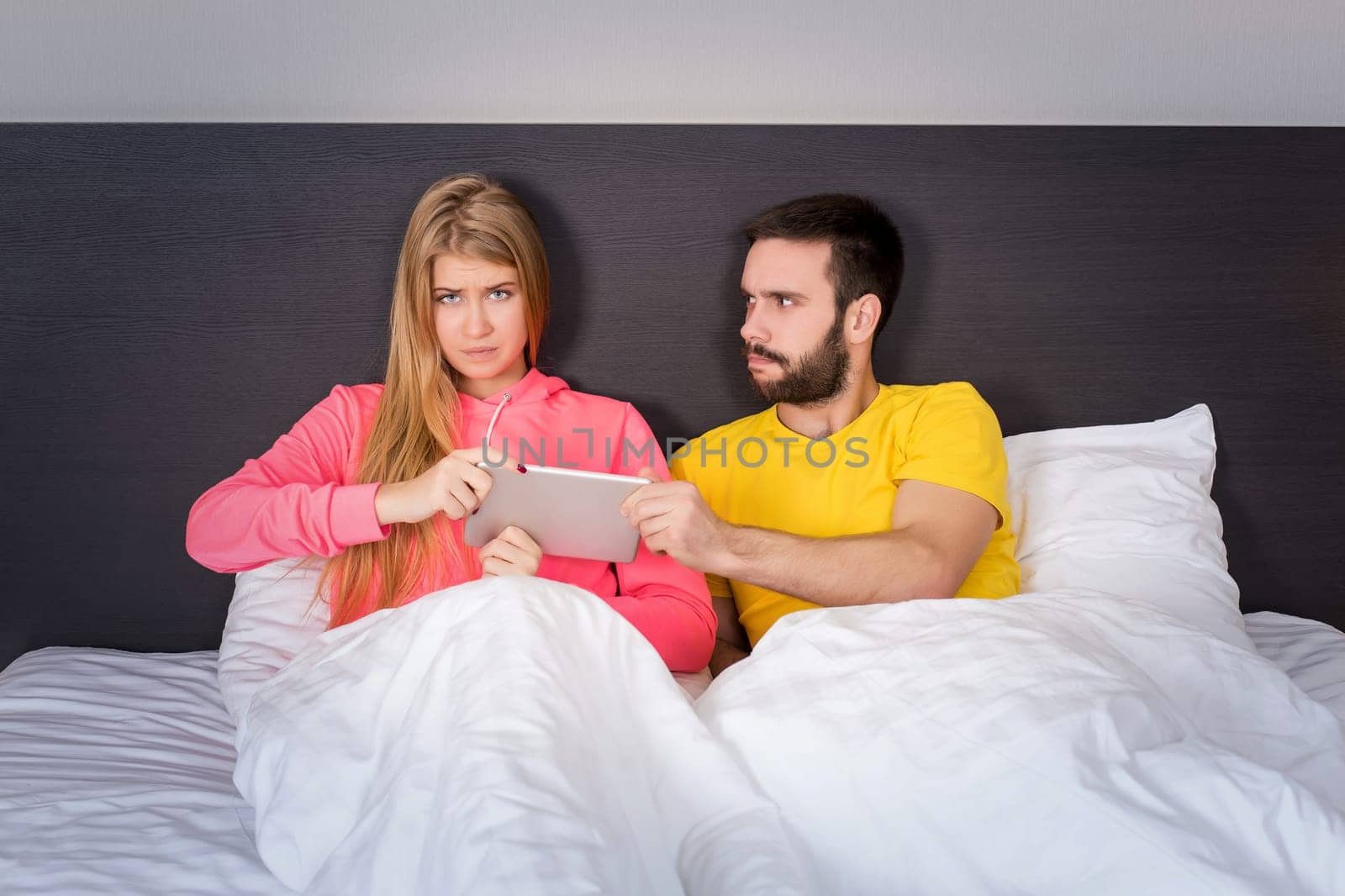Young sweet couple at bed with Tablet Gadget, man takes it girl. Concept about technology and people