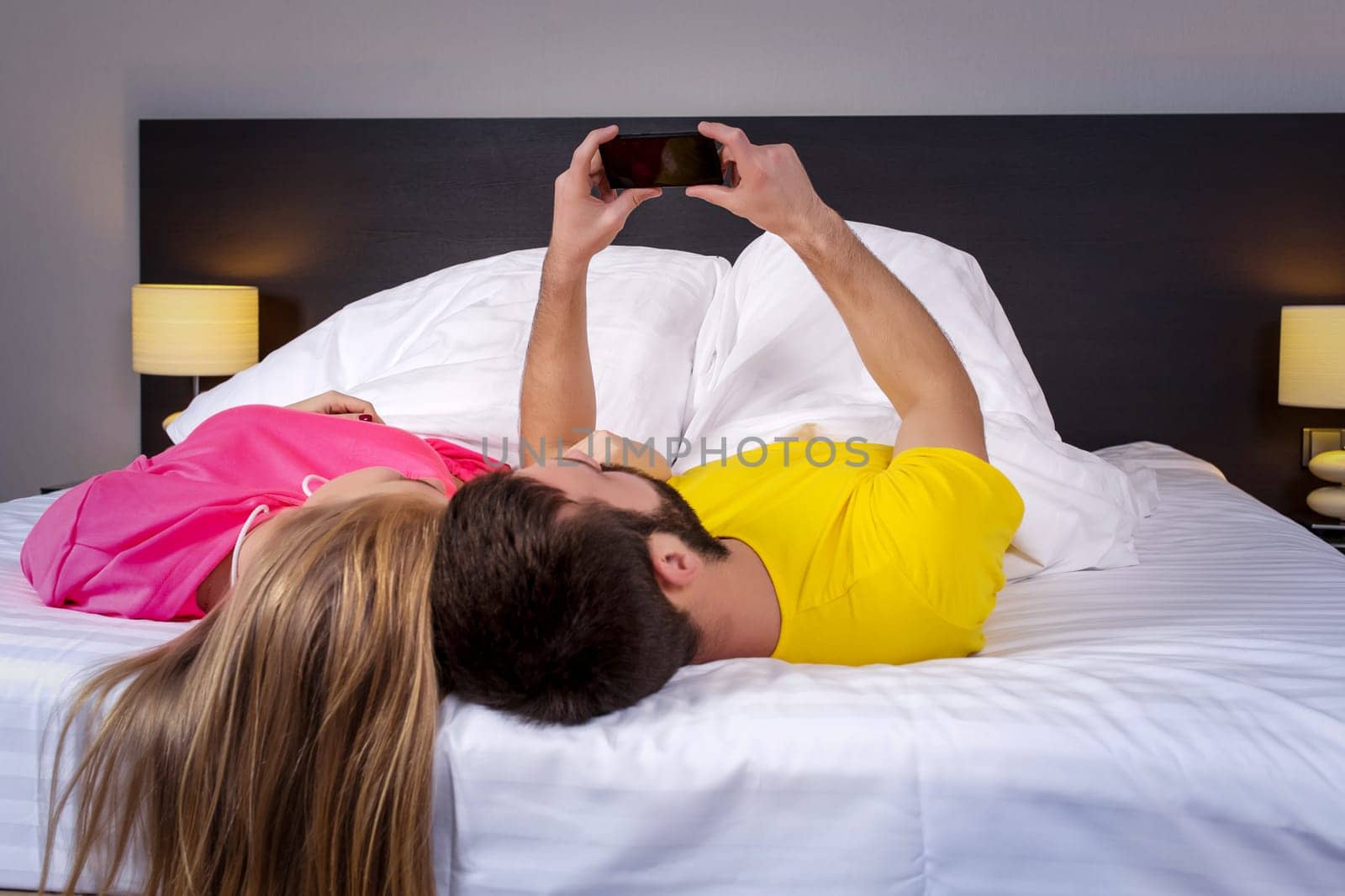 Young couple lying on their backs on the bed, the husband holding mobile phone and showing something to his wife