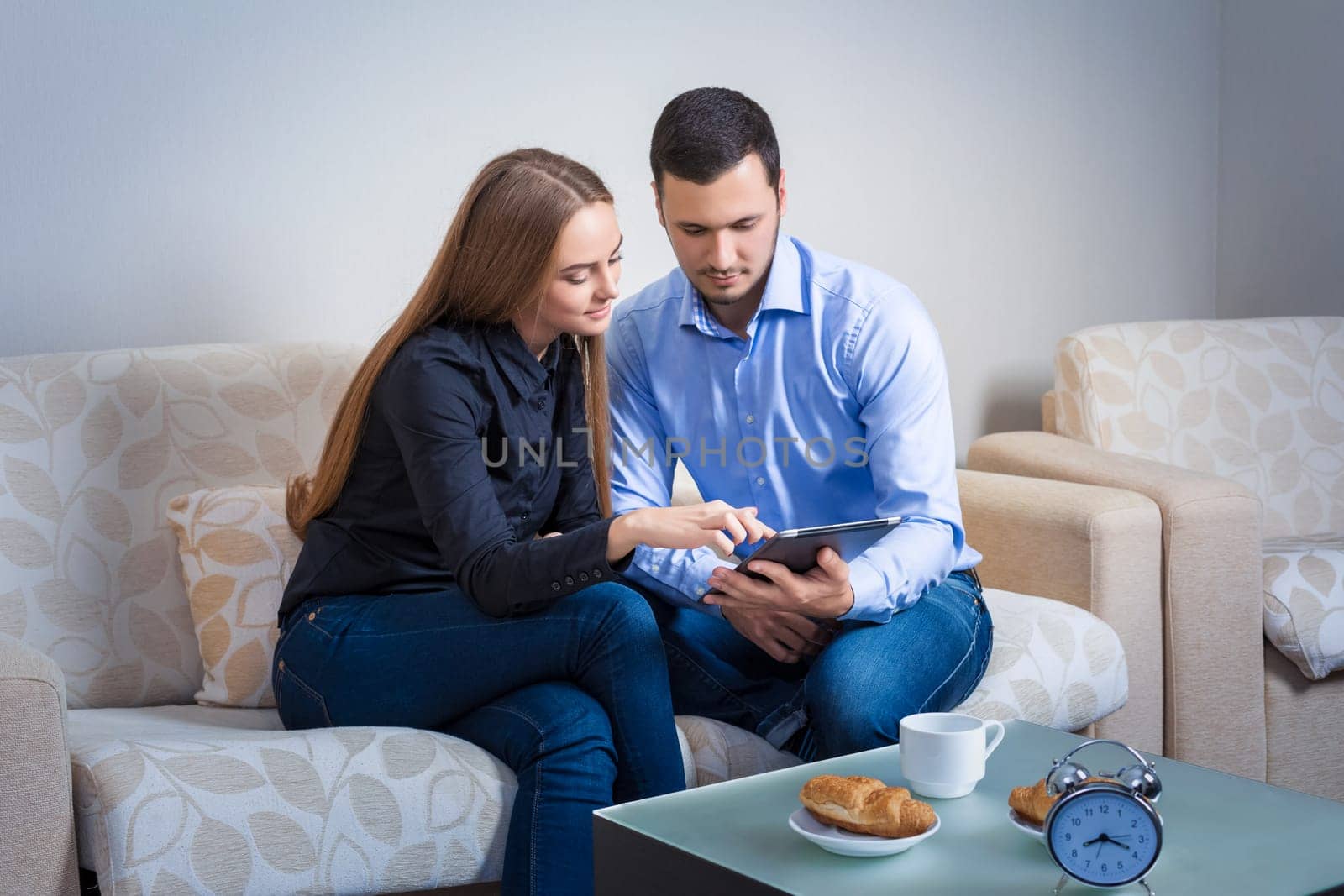 Young couple laughing, sitting on a sofa, sharing information, displayed on an electronic tablet with each other and drinking coffee