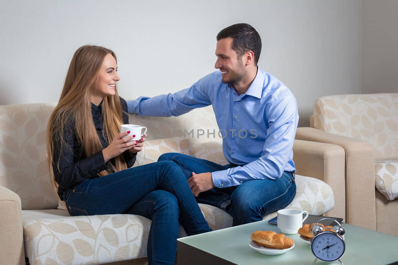 Beautiful young couple drink coffee with croissants, bvsitting on a sofa happy smile looking each other talking, portrait of lovely young man and woman on the couch