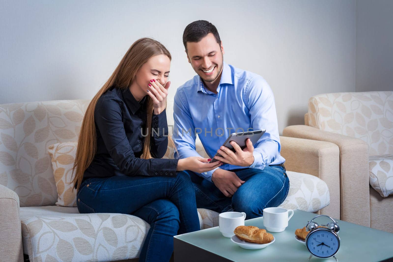 Young couple laughing, sitting on a sofa, sharing information, displayed on an electronic tablet with each other and drinking coffee