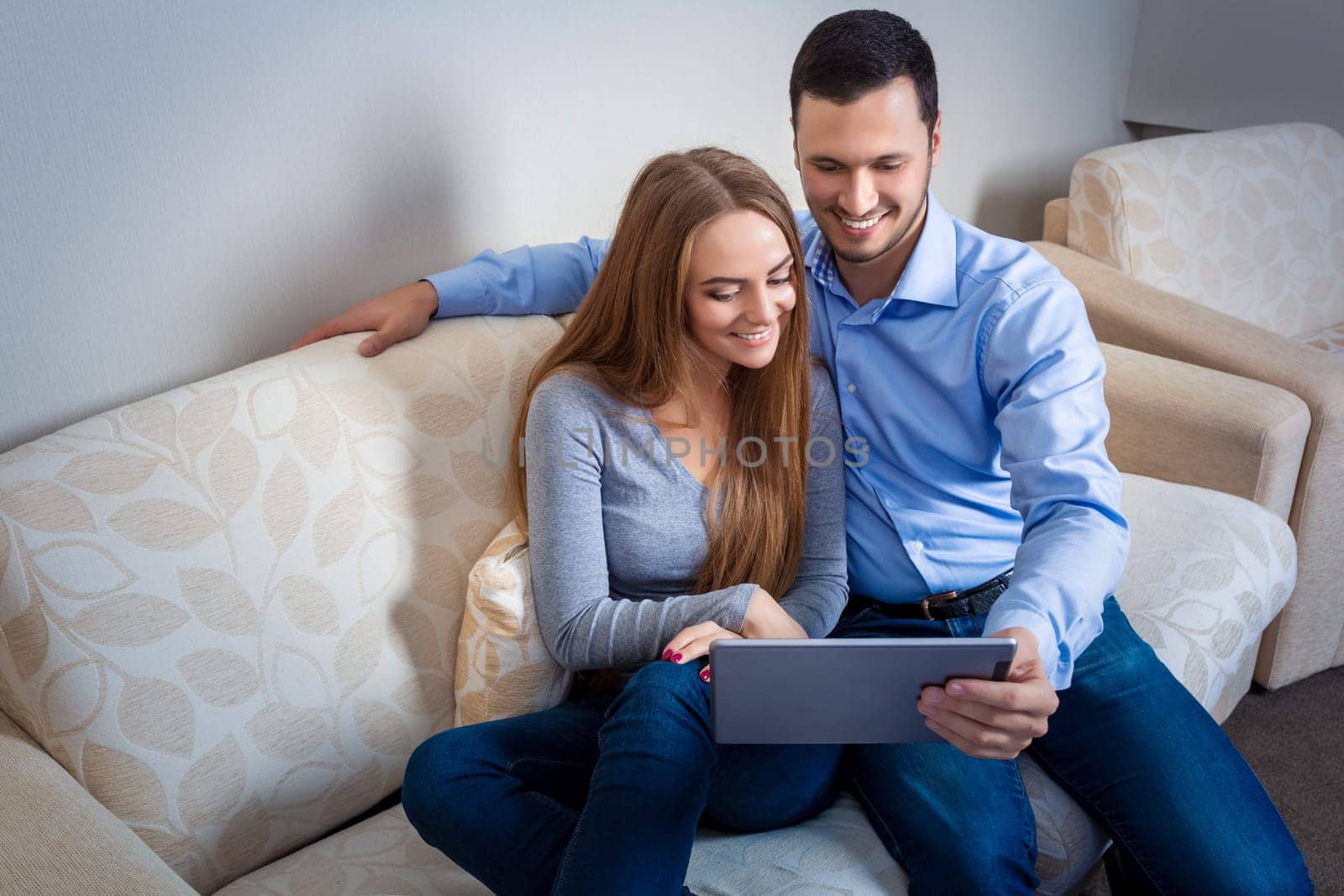 Beautiful young couple laughing, sitting on sofa, sharing photos or other information, displayed on an electronic tablet with each other