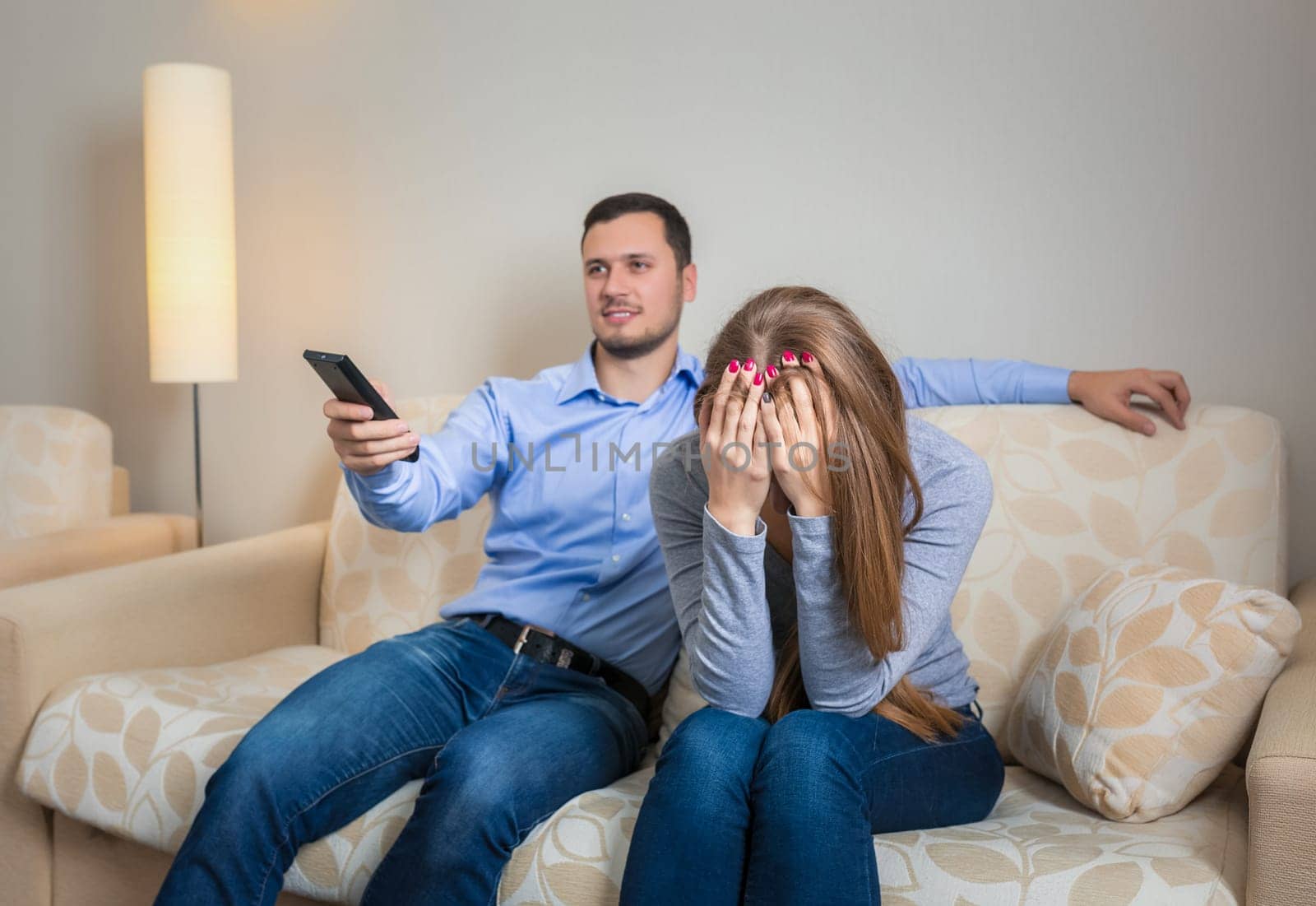 Portrait of couple sitting on sofa watching television. Image of men with remote control in hands and upset woman
