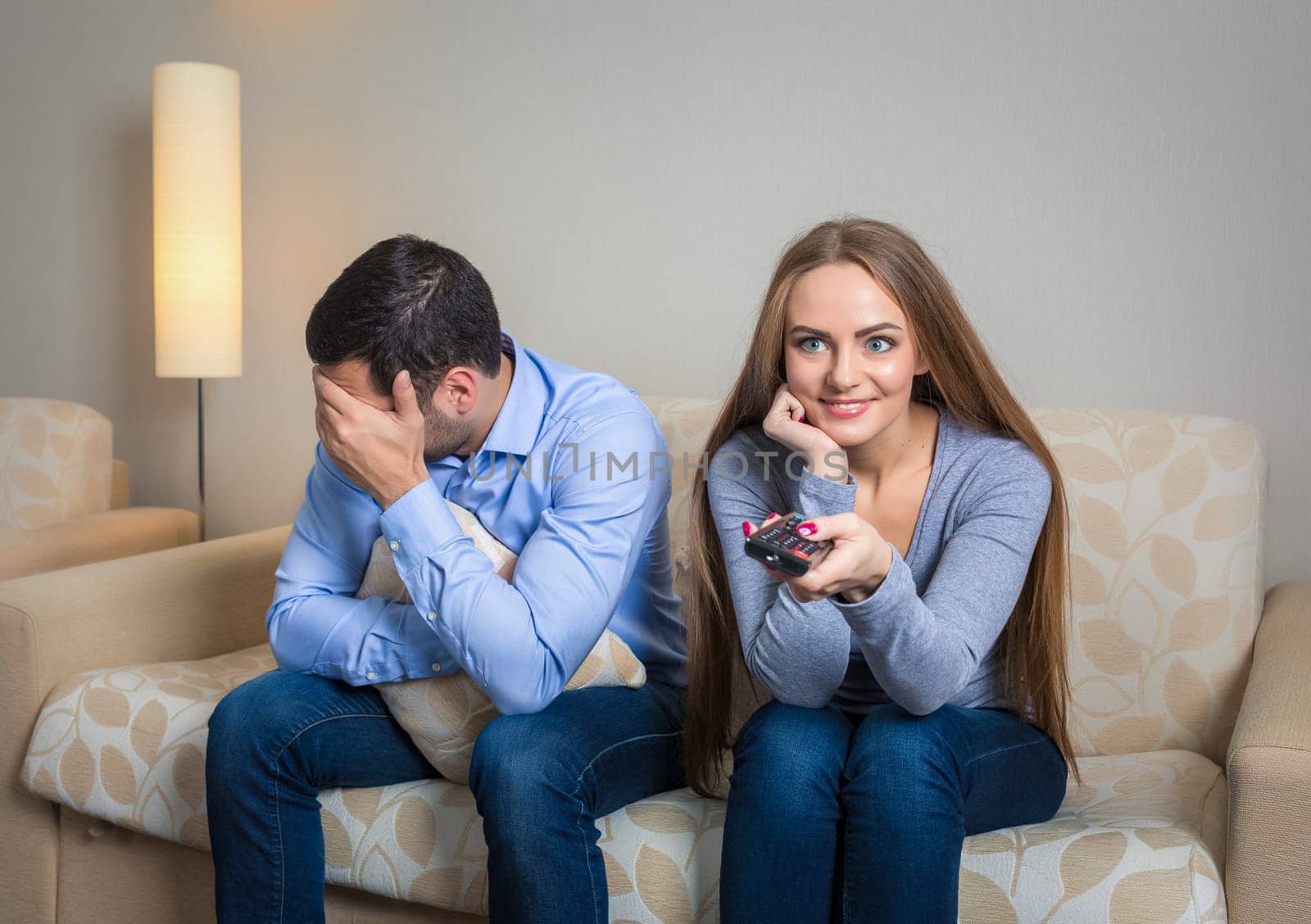Portrait of couple sitting on sofa watching television. Image of woman with remote control in hands and upset men
