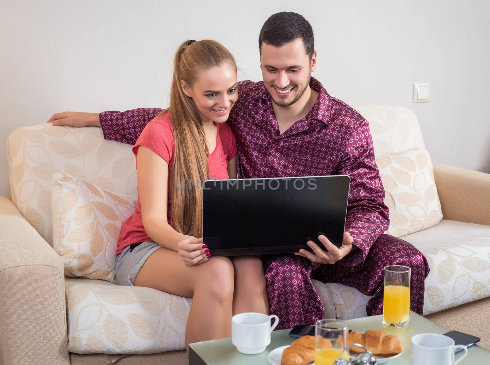Cute young couple on the couch having breakfast, eating croissants, drinking orange juice in front of a laptop computer