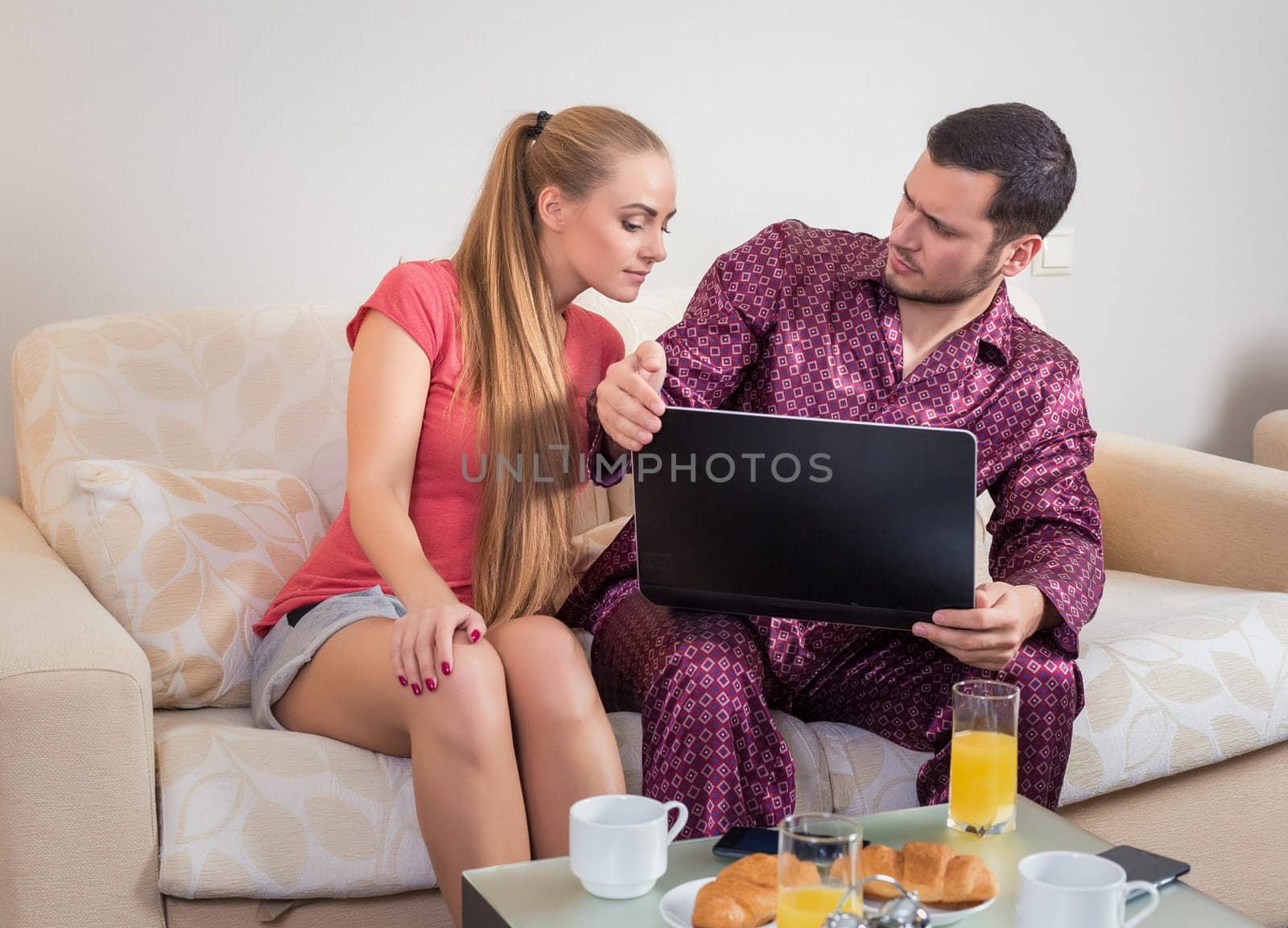 Cute young couple on the couch having breakfast, eating croissants, drinking orange juice in front of a laptop computer