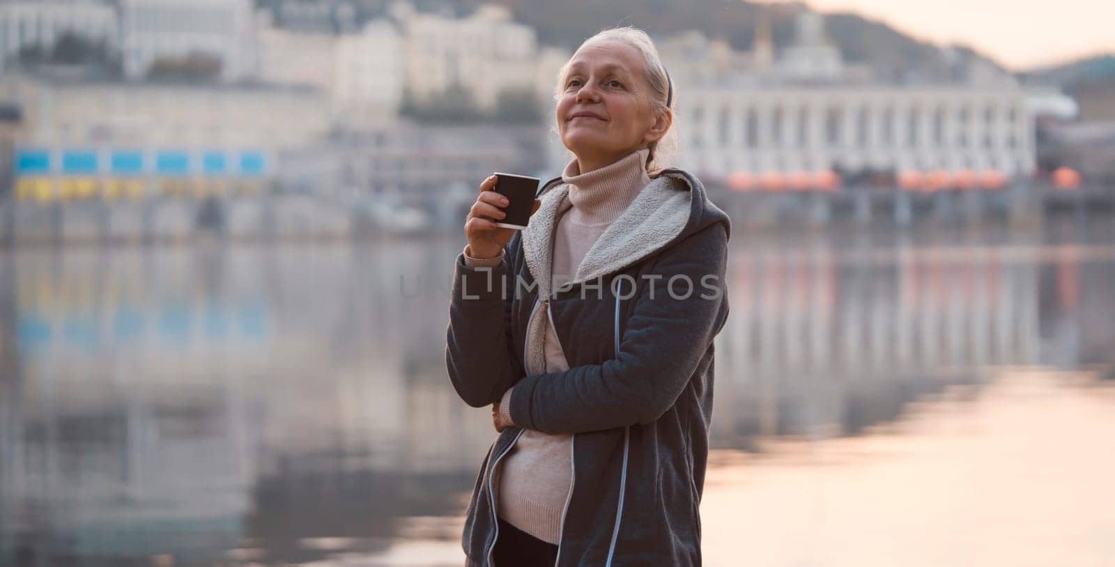 An adult beautiful woman holds a glass of aromatic coffee in her hands and enjoys the taste and atmosphere at sunset, views of the city and the river.