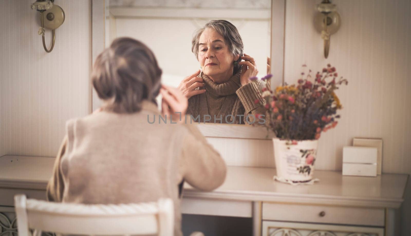 An elderly smiling woman of 80+ years of age spends a good time at home, a grandmother takes care of her appearance, looks in the mirror, sits at a dressing table in her bedroom.