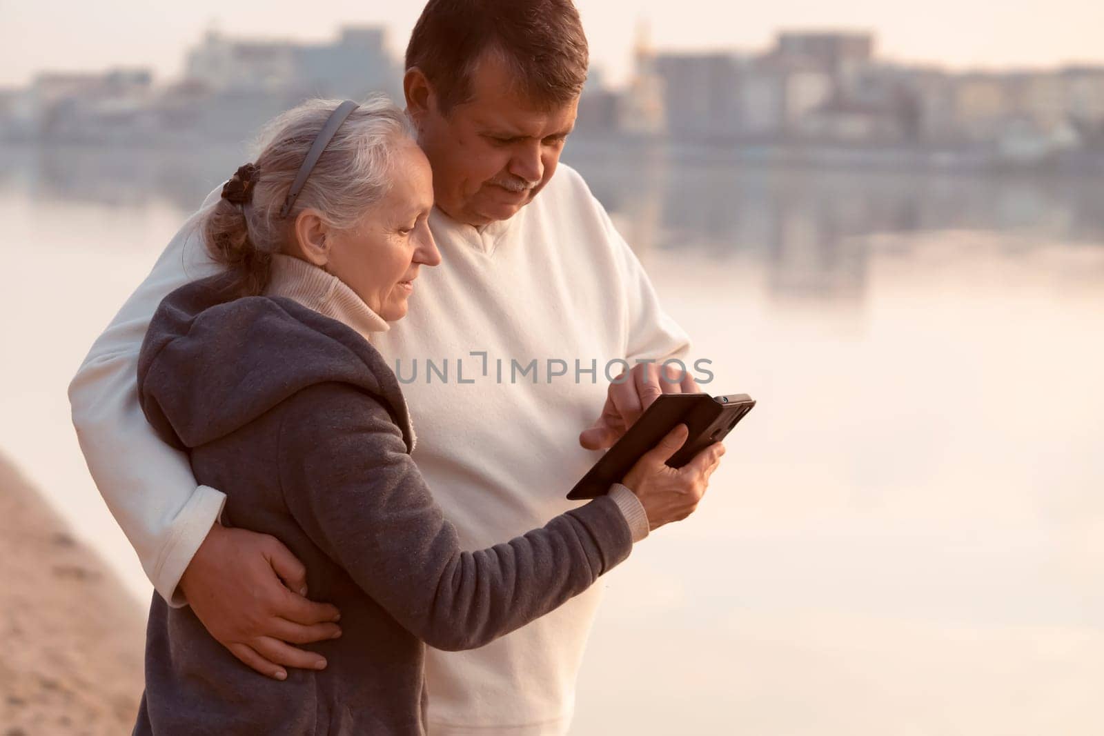 Family adult happy couple walks in the city park near the river at sunset, a gray-haired woman shows a photo on the phone to a man, husband and wife have a good leisure time together.