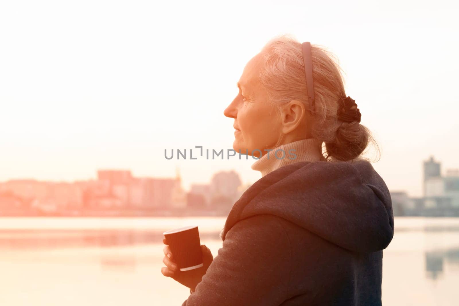 An adult beautiful woman holds a glass of aromatic coffee in her hands and enjoys the taste and atmosphere at sunset, views of the city and the river.