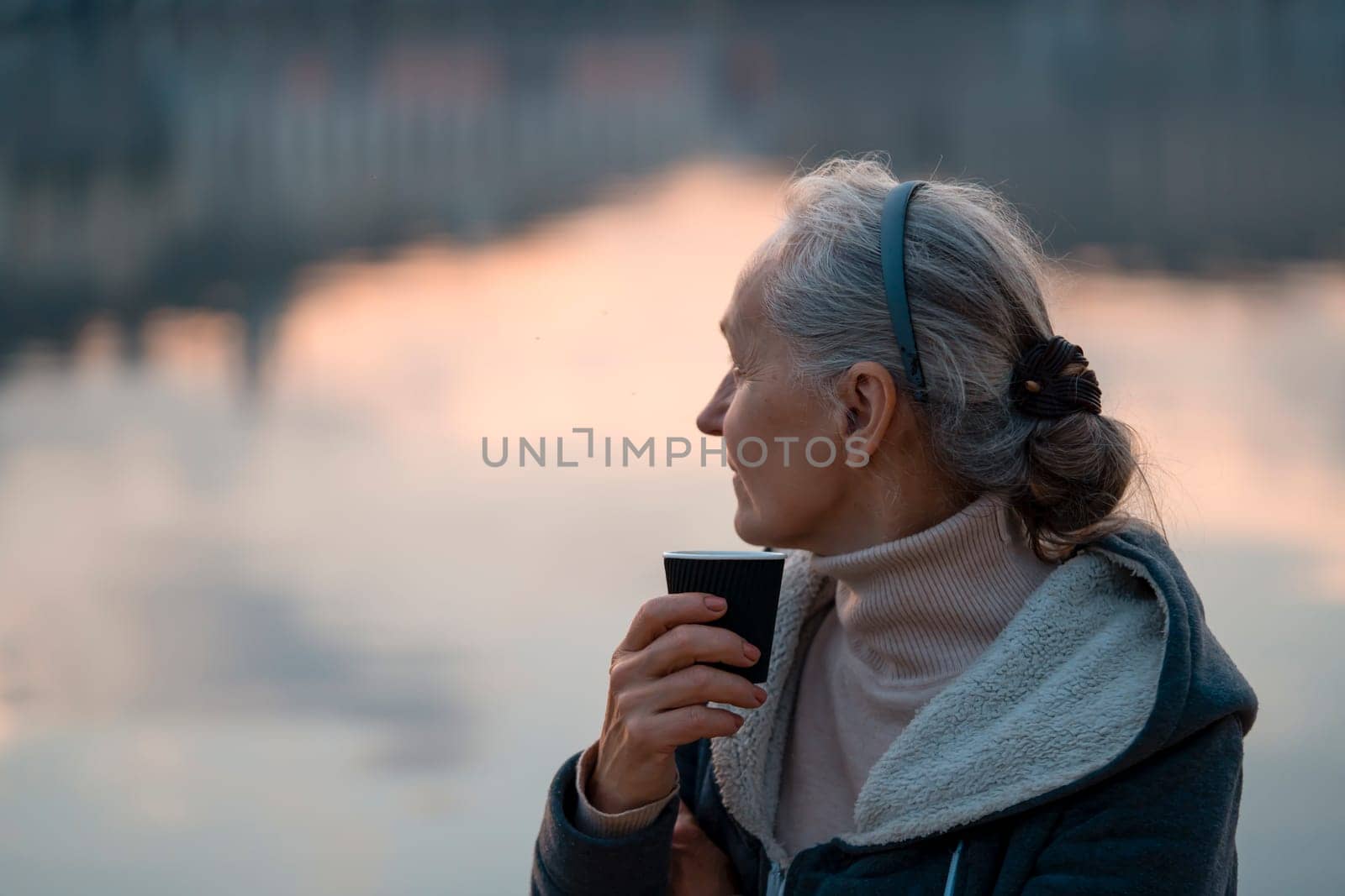 An adult beautiful woman holds a glass of aromatic coffee in her hands and enjoys the taste and atmosphere at sunset, views of the city and the river.