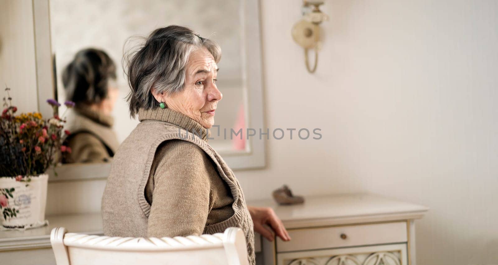 An elderly smiling woman of 80+ years of age spends a good time at home, a grandmother takes care of her appearance, looks in the mirror, sits at a dressing table in her bedroom.