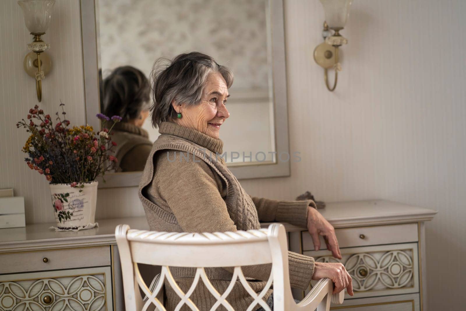 An elderly smiling woman of 80+ years of age spends a good time at home, a grandmother takes care of her appearance, looks in the mirror, sits at a dressing table in her bedroom.