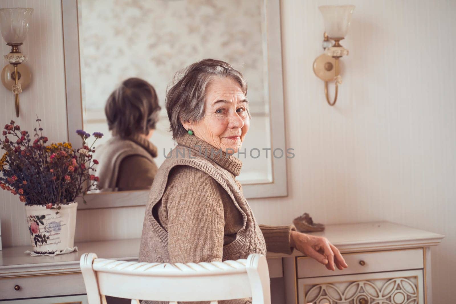 Beautiful old woman sits near the mirror in her cozy light room. by africapink