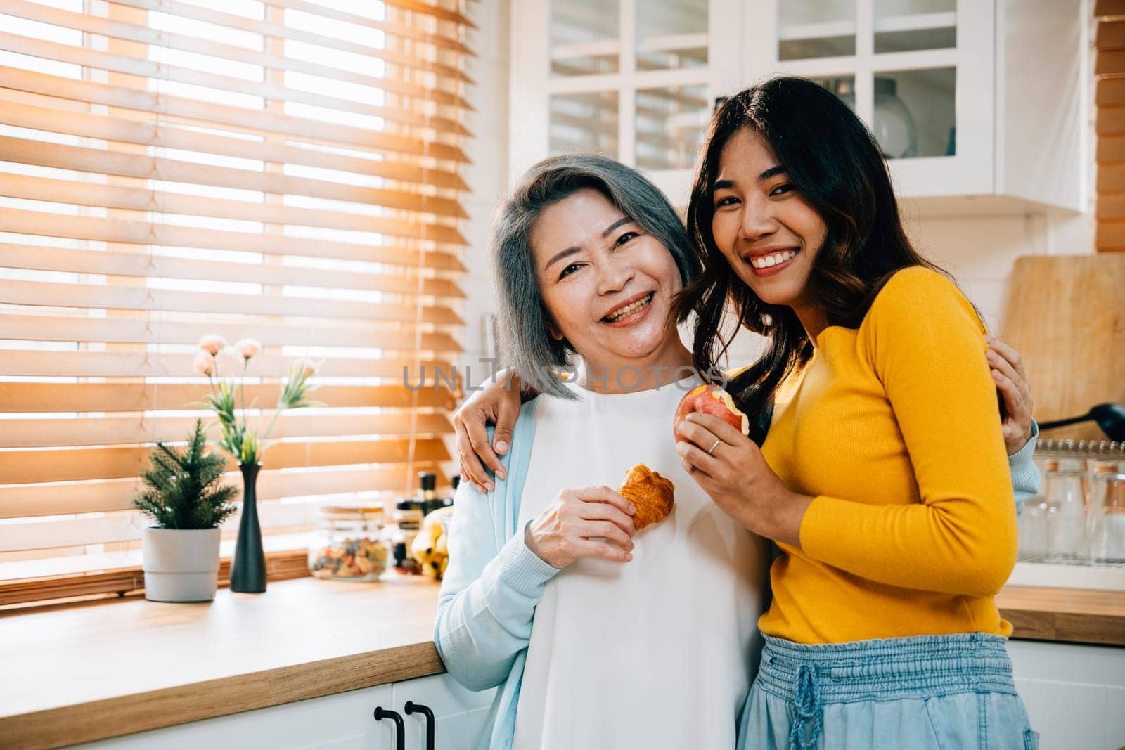 An elderly mother and her adult daughter, a cheerful young woman, hold an apple in the kitchen. Their smiles convey the joy of teaching, learning, and family togetherness. by Sorapop