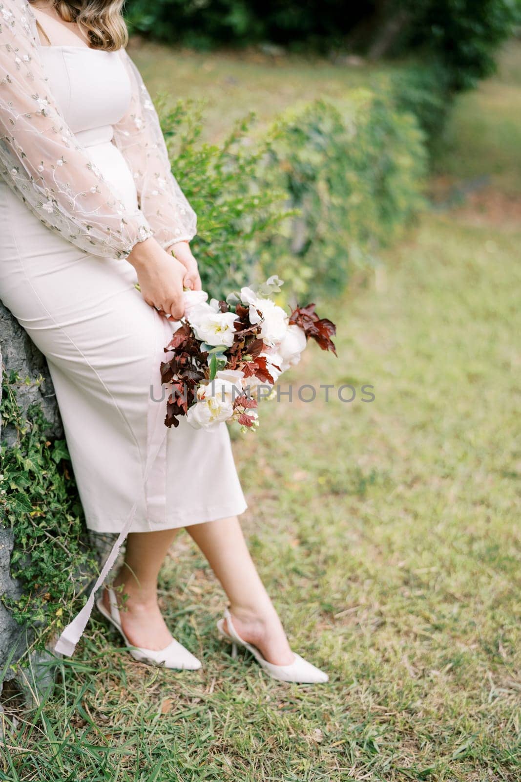 Bride with a bouquet in her hand is leaning against a hedge in the garden. Cropped. Faceless. High quality photo