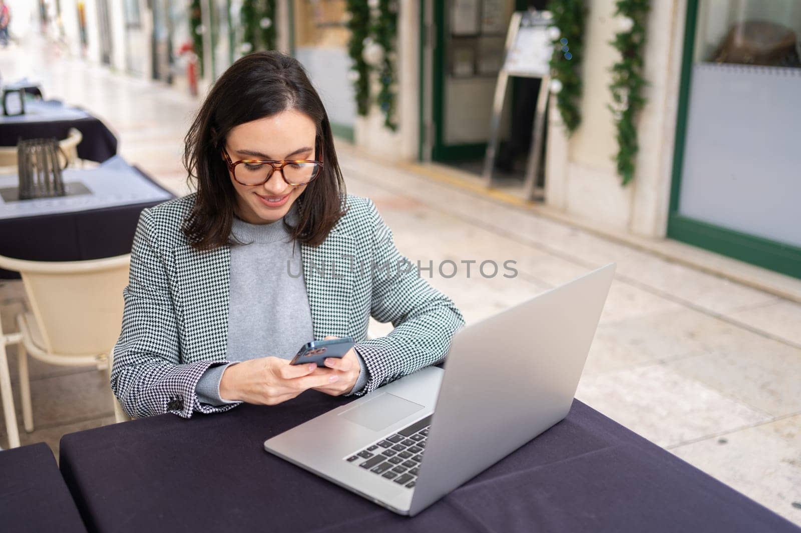 Freelance woman sitting in cafe working with laptop outdoor. Business woman in glasses and smart casual style, remotely working sitting in cafe with laptop and sending message use smartphone