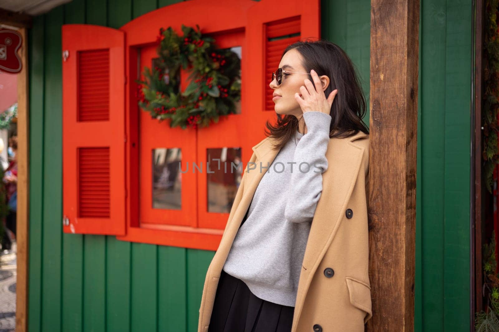 Woman standing sideways near Christmas store. Woman dressed stylish coat with sunglasses leaning on pole outside Christmas store. Female standing on street in fall wearing coat