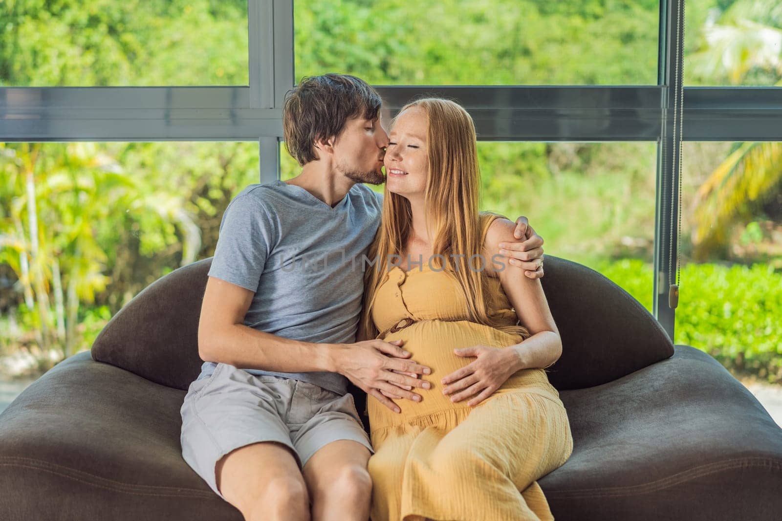 A tender moment captured as a husband and pregnant wife sit on the sofa, sharing an affectionate hug, their love and anticipation radiating in their embrace by galitskaya