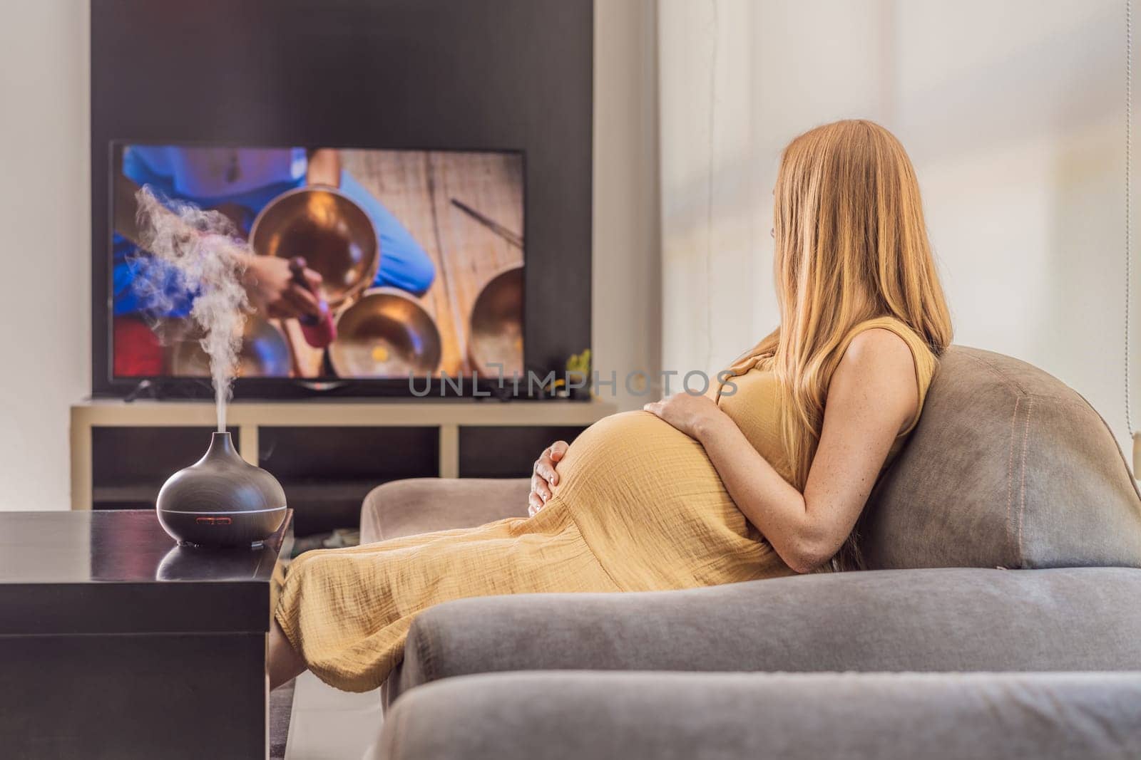 A blissful pregnant woman immerses in relaxation, savoring the soothing aroma from a diffuser while indulging in a calming TV video, embracing tranquility during her pregnant journey.