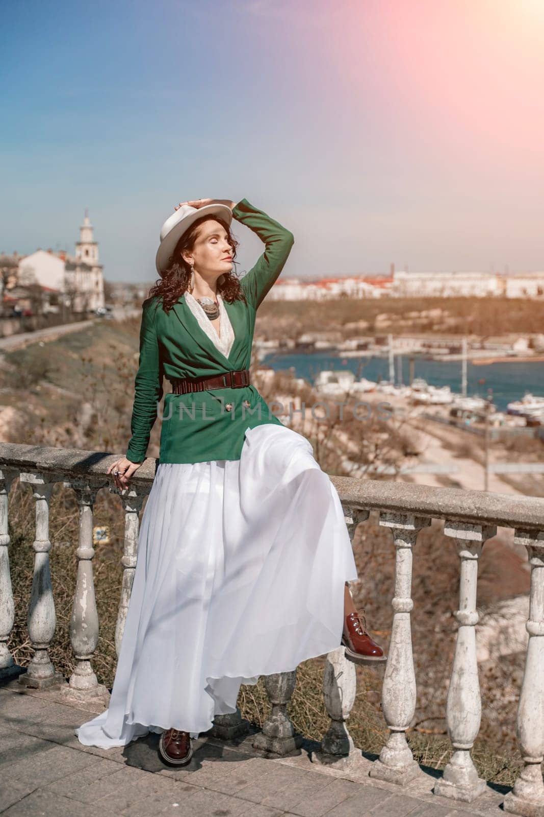 Woman walks around the city, lifestyle. A young beautiful woman in a green jacket, white skirt and hat is sitting on a white fence with balusters overlooking the sea bay and the city. by Matiunina