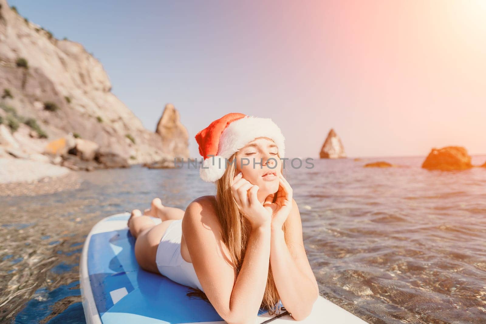 Woman sea sup. Close up portrait of happy young caucasian woman with long hair in Santa hat looking at camera and smiling. Cute woman portrait in a white bikini posing on sup board in the sea by panophotograph