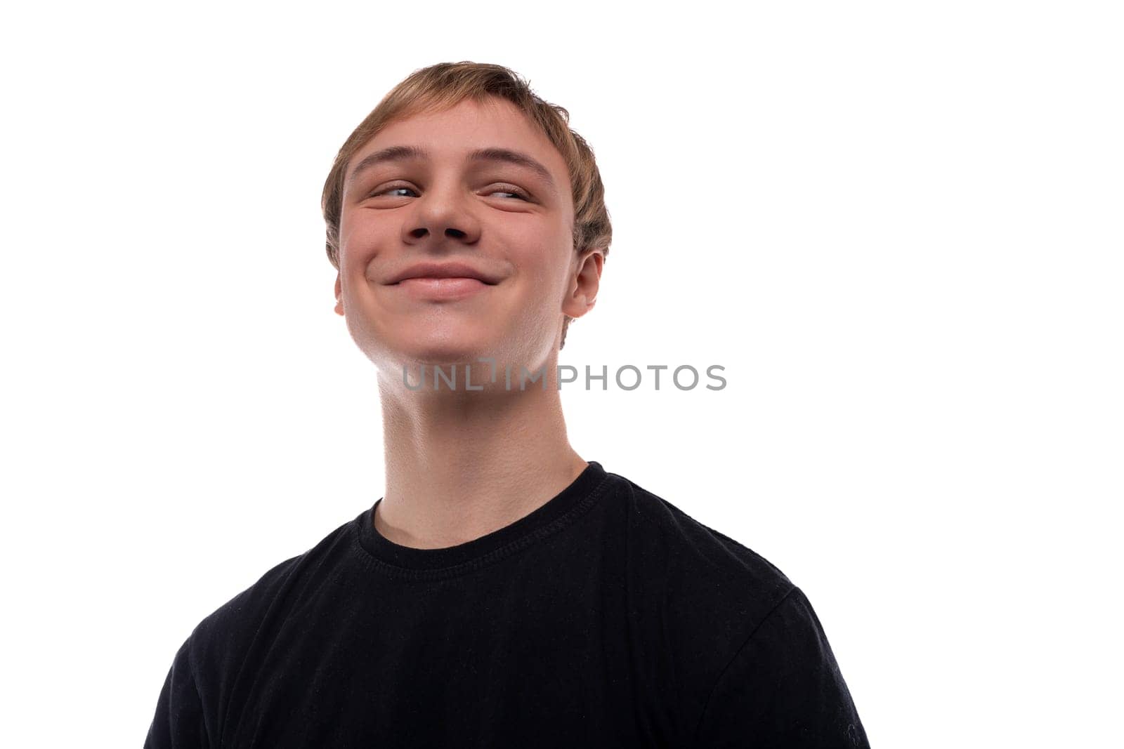 Handsome Caucasian blond teenager guy in a black T-shirt smiles cutely and looks to the side.