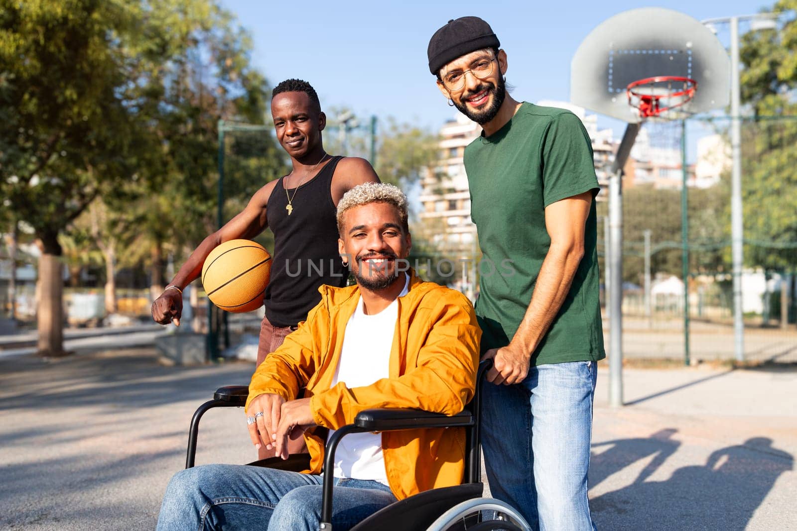 Happy young African American man in a wheelchair with friends in basketball court outdoors looking at camera smiling. Sports and friendship concept.