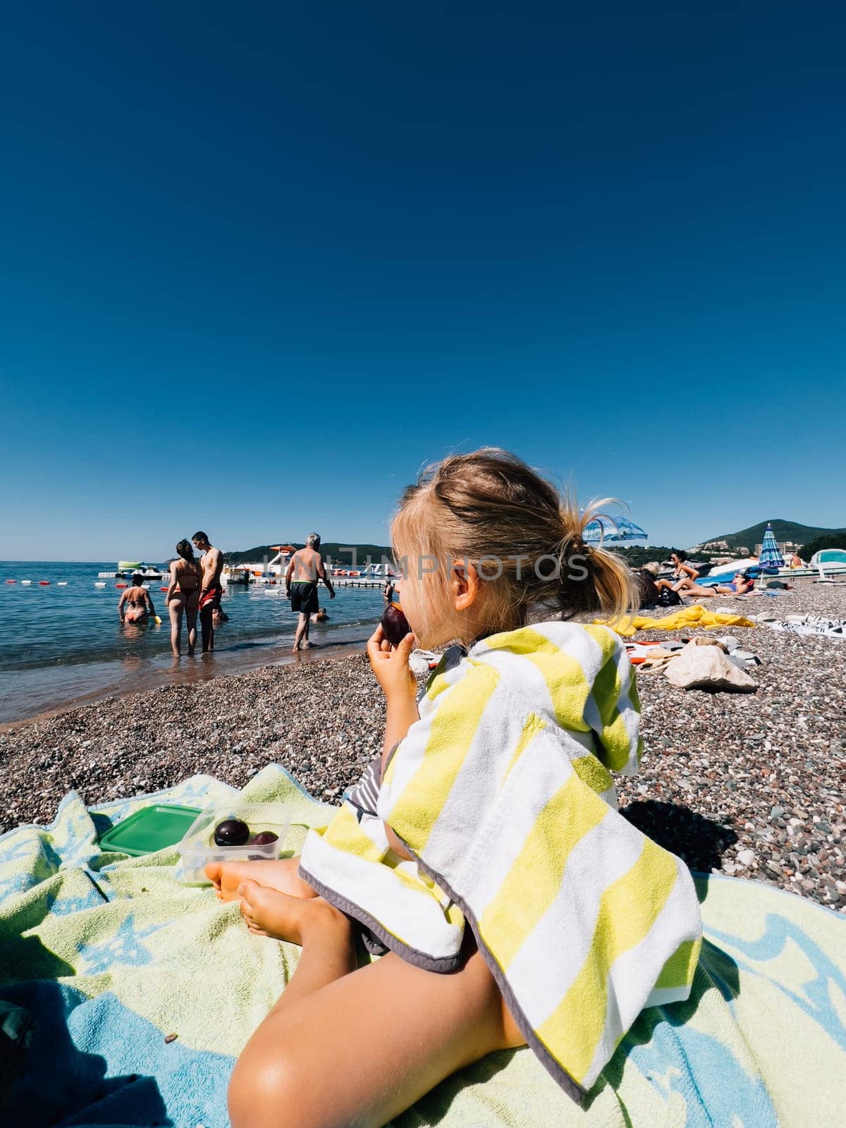 Becici, Montenegro - 03 august 2023: Little girl sits in a towel on the beach and looks at the sea by Nadtochiy