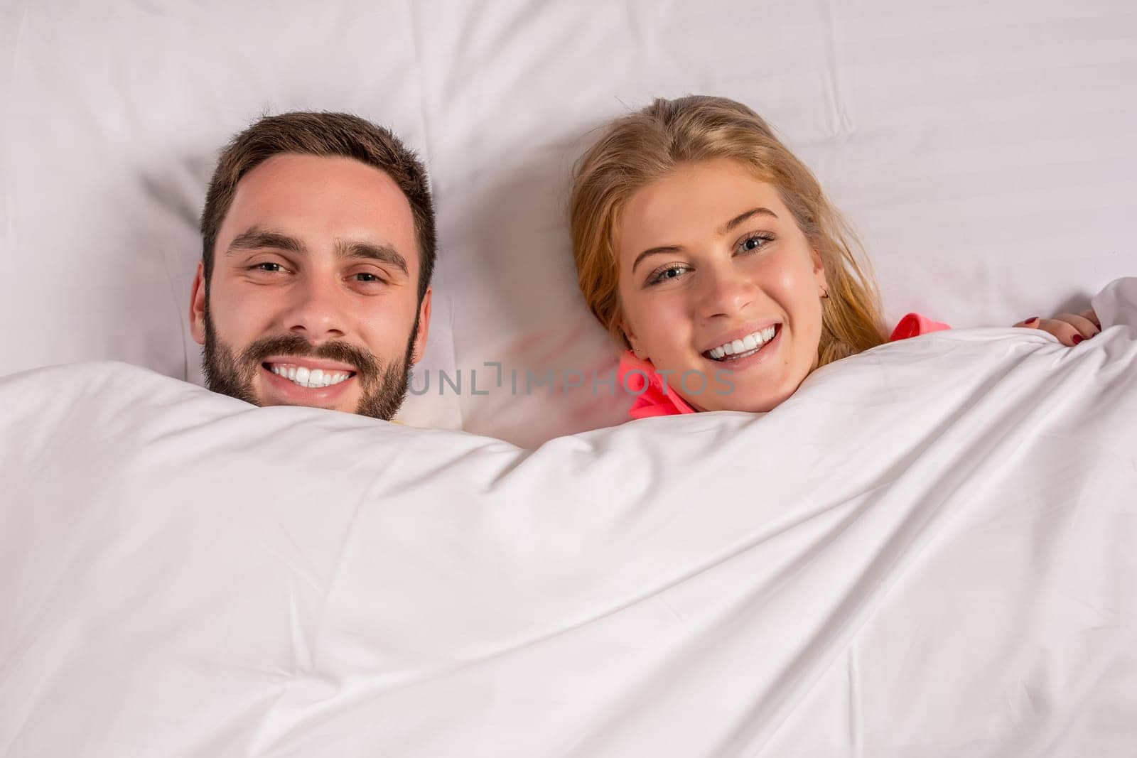 Young lovely couple lying and have fun in a bed, happy smile looking at camera, cover under blanket. Family, bedtime and happiness concept