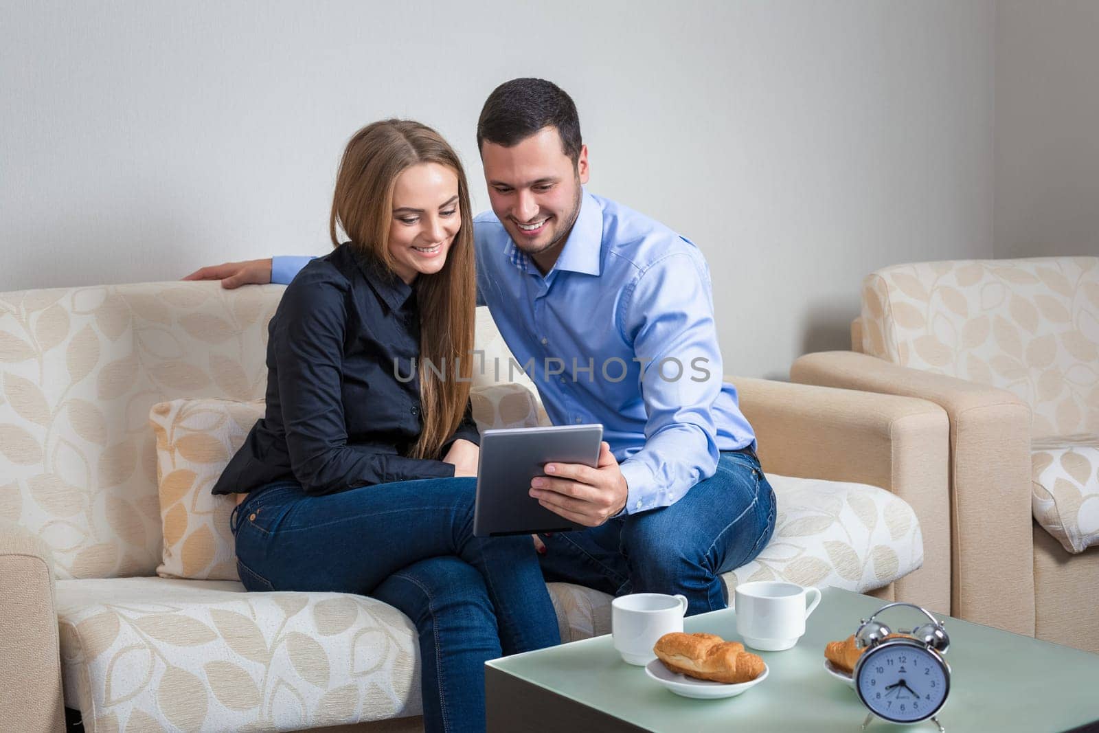 Beautiful young couple laughing, bvsitting on a sofa, sharing photos or other information, displayed on an electronic tablet with each other and drinking coffee with croissants