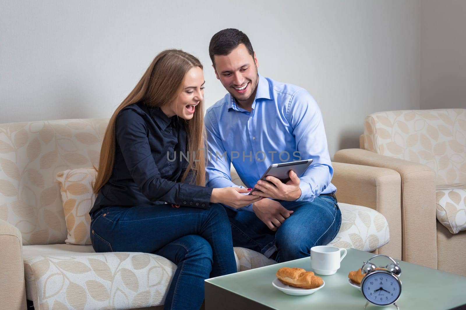 Beautiful young couple laughing, bvsitting on a sofa, sharing photos or other information, displayed on an electronic tablet with each other and drinking coffee with croissants