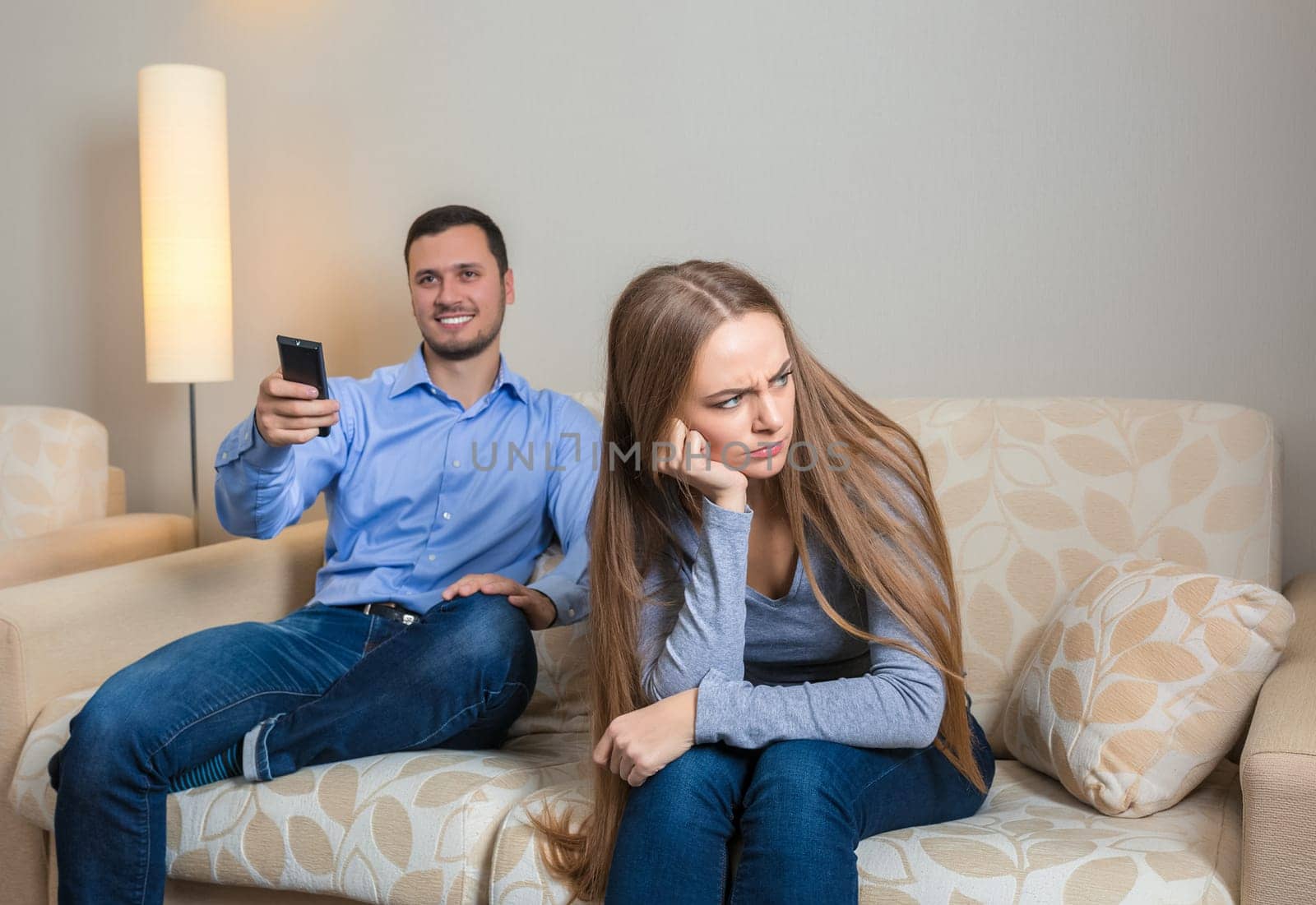 Portrait of couple sitting on sofa watching television. Image of men with remote control in hands and upset woman