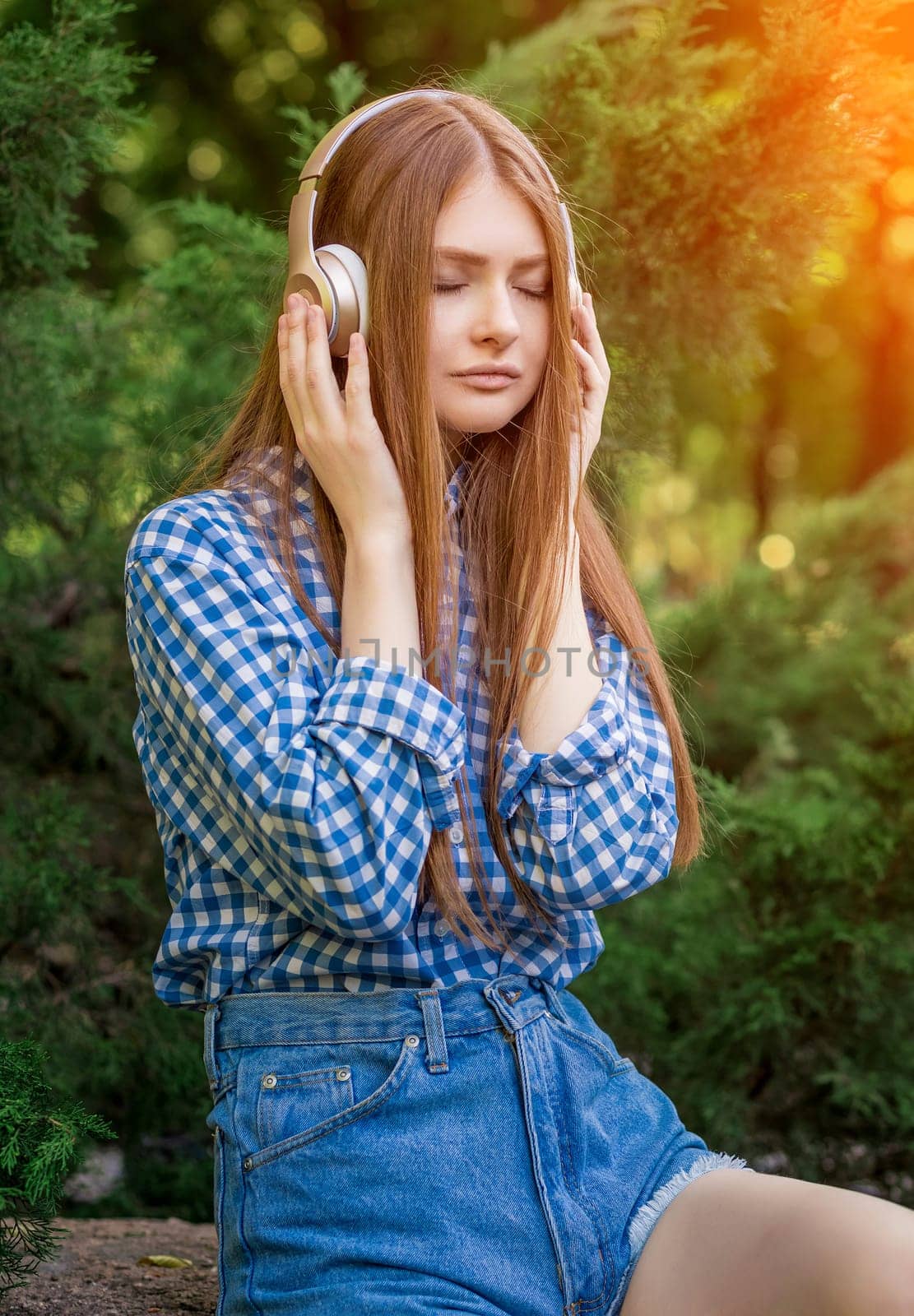Young woman cute brunette girl sitting on stone on headphones with eyes closed, sun light flares of rays, on summer outdoor copy space background